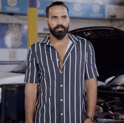 A Man With A Beard Wearing A Striped Shirt Stands In Front Of A Car GIF