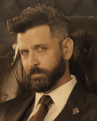 A Man With A Beard Wearing A Suit And Tie Is Sitting In A Chair GIF