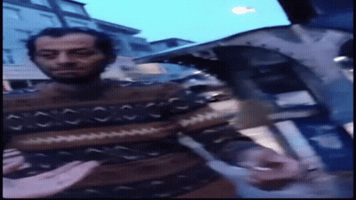 A Man With A Beard Wearing A Sweater Is Standing On A City Street GIF