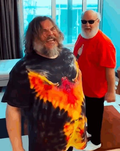A Man With A Beard Wearing A Tie Dye Shirt Stands Next To Another Man Wearing A Red Shirt GIF