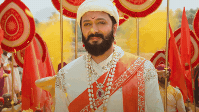 A Man With A Beard Wearing A Turban And Pearls Stands In Front Of Red Flags GIF