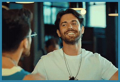 A Man With A Beard Wearing A White T-Shirt Smiles While Talking To Another Man GIF