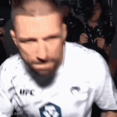 A Man With A Beard Wearing A White Ufc Shirt Is Standing In Front Of A Crowd . GIF