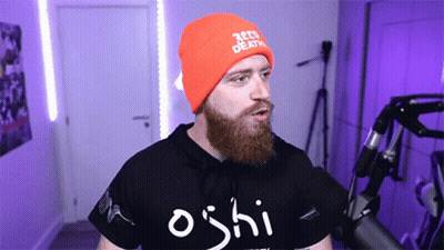 A Man With A Beard Wearing An Orange Beanie And A Black Shirt That Says Oshi GIF