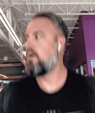 A Man With A Beard Wearing Ear Buds And A Black Shirt With A Purple Wall In The Background GIF