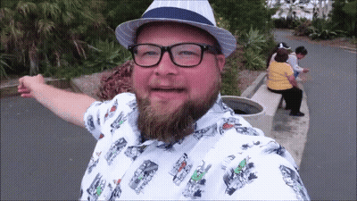 A Man With A Beard Wearing Glasses And A Hat With A Shirt With Cars On It GIF