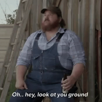 A Man With A Beard Wearing Overalls And A Plaid Shirt Is Sitting On A Ladder Holding A Beer . GIF