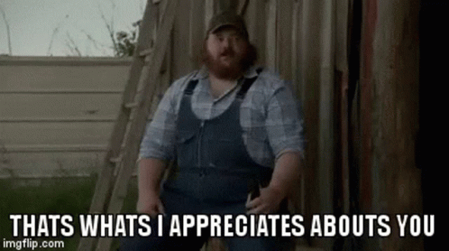 A Man With A Beard Wearing Overalls Is Sitting In Front Of A Wooden Shed . GIF