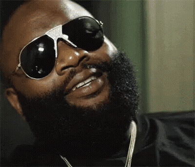 A Man With A Beard Wearing Sunglasses And A Necklace Is Smiling . GIF