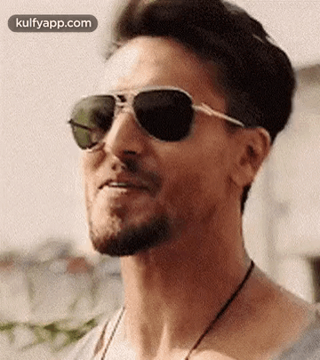 A Man With A Beard Wearing Sunglasses And A Necklace Is Smiling . GIF