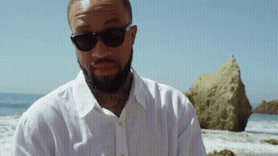 A Man With A Beard Wearing Sunglasses And A White Shirt Stands On The Beach GIF