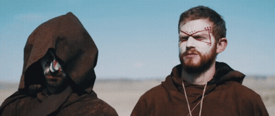 A Man With A Cross Painted On His Face Stands Next To Another Man With A Hood GIF