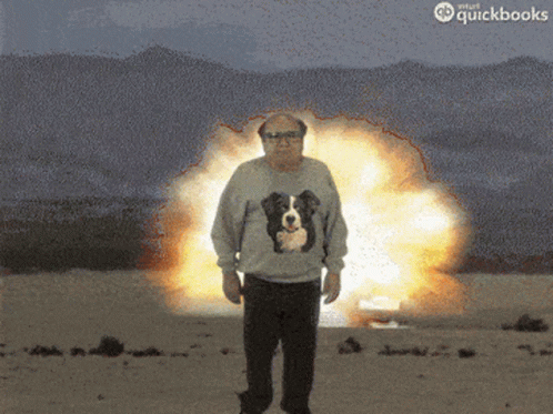 A Man With A Dog On His Sweater Stands In Front Of An Explosion GIF