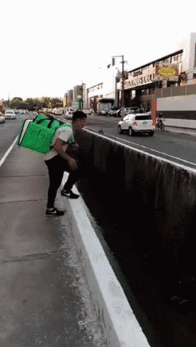 A Man With A Green Bag On His Back Is Standing On A Sidewalk Next To A Street GIF
