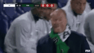 A Man With A Green Scarf Around His Neck Is Sitting In The Stands Watching A Soccer Game . GIF