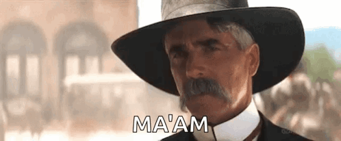 A Man With A Hat And Mustache Is Standing In Front Of A Building And Saying Ma 'Am . GIF