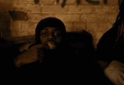 A Man With A Hood On Looks At The Camera With Graffiti On The Wall Behind Him GIF
