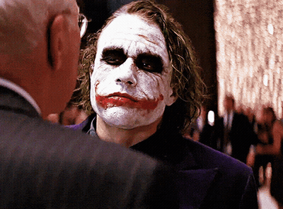 A Man With A Joker Face Painted On His Face Talks To Another Man In A Suit GIF