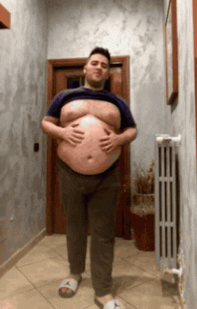 A Man With A Large Belly Is Standing In A Hallway Holding His Belly GIF