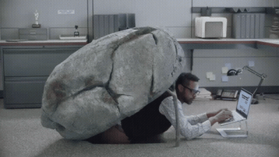 A Man With A Large Rock On His Back Is Using A Laptop In An Office GIF