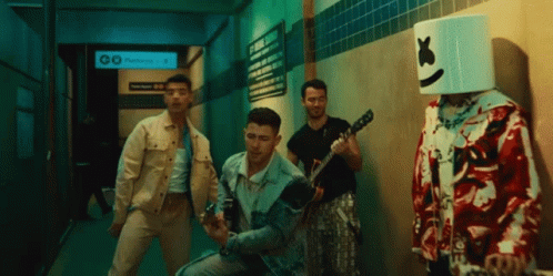 A Man With A Marshmallow Head Stands Next To A Group Of Men Playing Guitars GIF