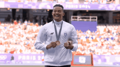 A Man With A Medal Around His Neck Stands In Front Of A Stadium That Says Paris 2021 GIF