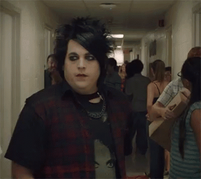 A Man With A Mohawk And Black Eye Makeup Is Standing In A Hallway Surrounded By People . GIF