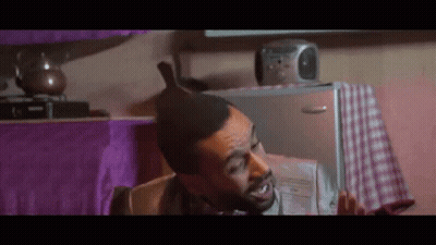 A Man With A Mohawk Is Sitting In Front Of A Refrigerator Holding A Newspaper . GIF