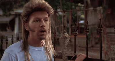 A Man With A Mullet And A Beard Is Standing In Front Of A Metal Fence GIF