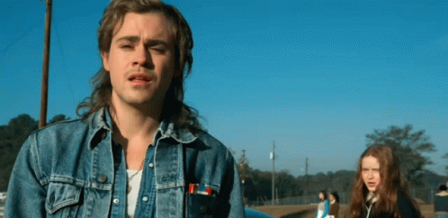A Man With A Mullet And A Denim Jacket Is Standing In Front Of A Blue Car . GIF