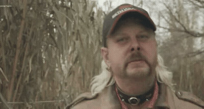 A Man With A Mullet And A Hat Is Standing In Front Of A Fence In A Field . GIF