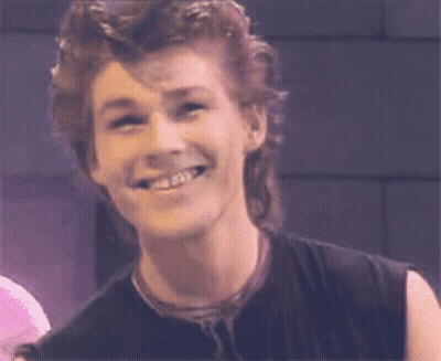 A Man With A Mullet Is Smiling And Wearing A Black Shirt GIF