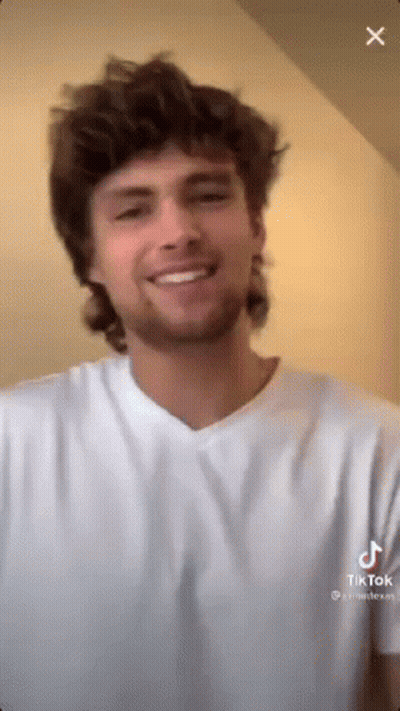 A Man With A Mullet Is Smiling And Wearing A White Shirt . GIF