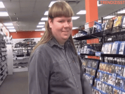 A Man With A Mullet Is Standing In Front Of A Shelf Of Dvds GIF