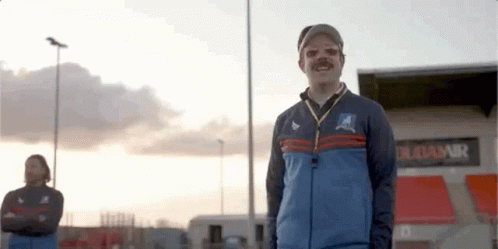 A Man With A Mustache And A Hat Is Standing In Front Of A Stadium . GIF