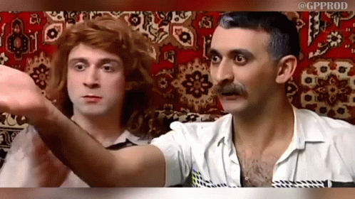 A Man With A Mustache And A Woman With Red Hair Are Sitting Next To Each Other On A Rug GIF