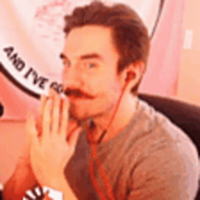 A Man With A Mustache And Ear Buds Is Sitting In Front Of A Pink Sign . GIF