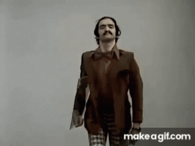 A Man With A Mustache And Headphones Is Standing Next To A Woman In A Plaid Shirt . GIF