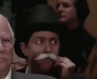 A Man With A Mustache And Top Hat Is Sitting In A Crowd Of People . GIF