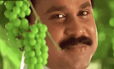 A Man With A Mustache Is Holding A Bunch Of Grapes Over His Face . GIF