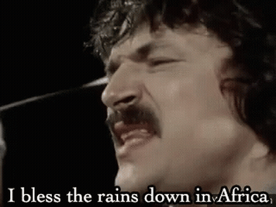 A Man With A Mustache Is Singing Into A Microphone And Saying `` I Bless The Rain Down In Africa '' . GIF