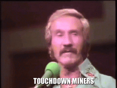 A Man With A Mustache Is Singing Into A Microphone With The Words Touchdown Miners Written On The Screen Behind Him . GIF