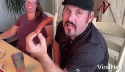 A Man With A Mustache Is Sitting At A Table With A Woman Holding A Hot Dog . GIF