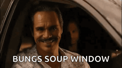 A Man With A Mustache Is Sitting In A Car With The Words Bungs Soup Window Written On The Screen Behind Him . GIF