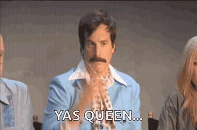 A Man With A Mustache Is Sitting In Front Of A Group Of People And Saying Yas Queen GIF