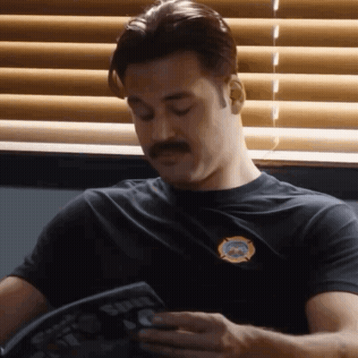 A Man With A Mustache Is Sitting In Front Of A Window Reading A Book . GIF