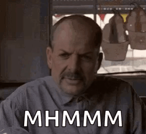 A Man With A Mustache Is Sitting In Front Of A Window With A Sign That Says Mhmm . GIF