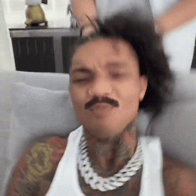 A Man With A Mustache Is Sitting On A Couch With His Hair In A Ponytail . GIF