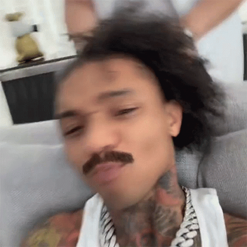 A Man With A Mustache Is Sitting On A Couch With His Hair In A Ponytail . GIF