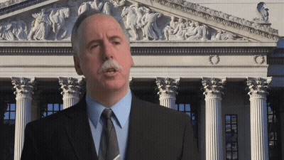 A Man With A Mustache Is Standing In Front Of A Building With Columns GIF